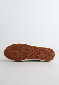 Podeszwa sportowego buta, z teksturowaną, gumową brązową powierzchnią o wzorku zapewniającym przyczepność. Krawędź jest biała z gładkim wykończeniem.