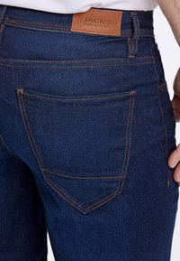 Jean en denim d'une teinte bleu foncé, avec des coutures orange, une poche arrière au design incurvé et une étiquette en cuir marron à la taille.