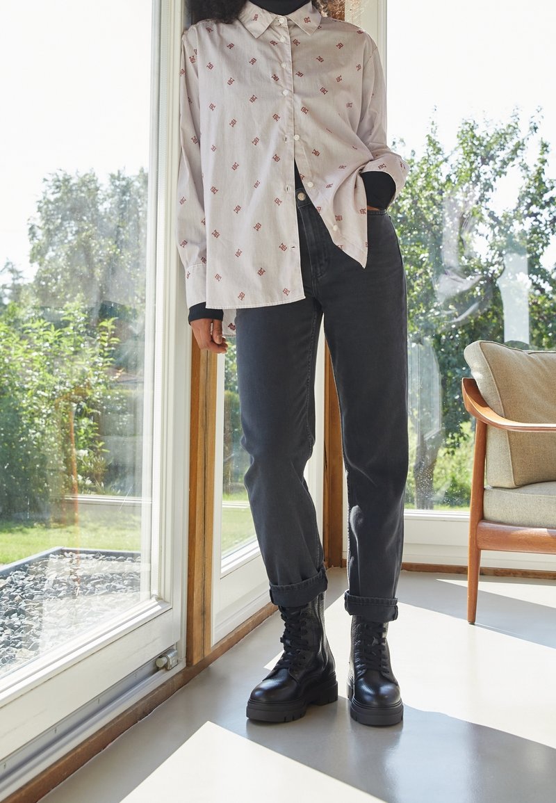 Personne portant une chemise blanche à motifs boutonnée, un jean noir retroussé et des bottes noires à lacets, debout à l'intérieur près d'une porte vitrée avec la lumière du soleil.