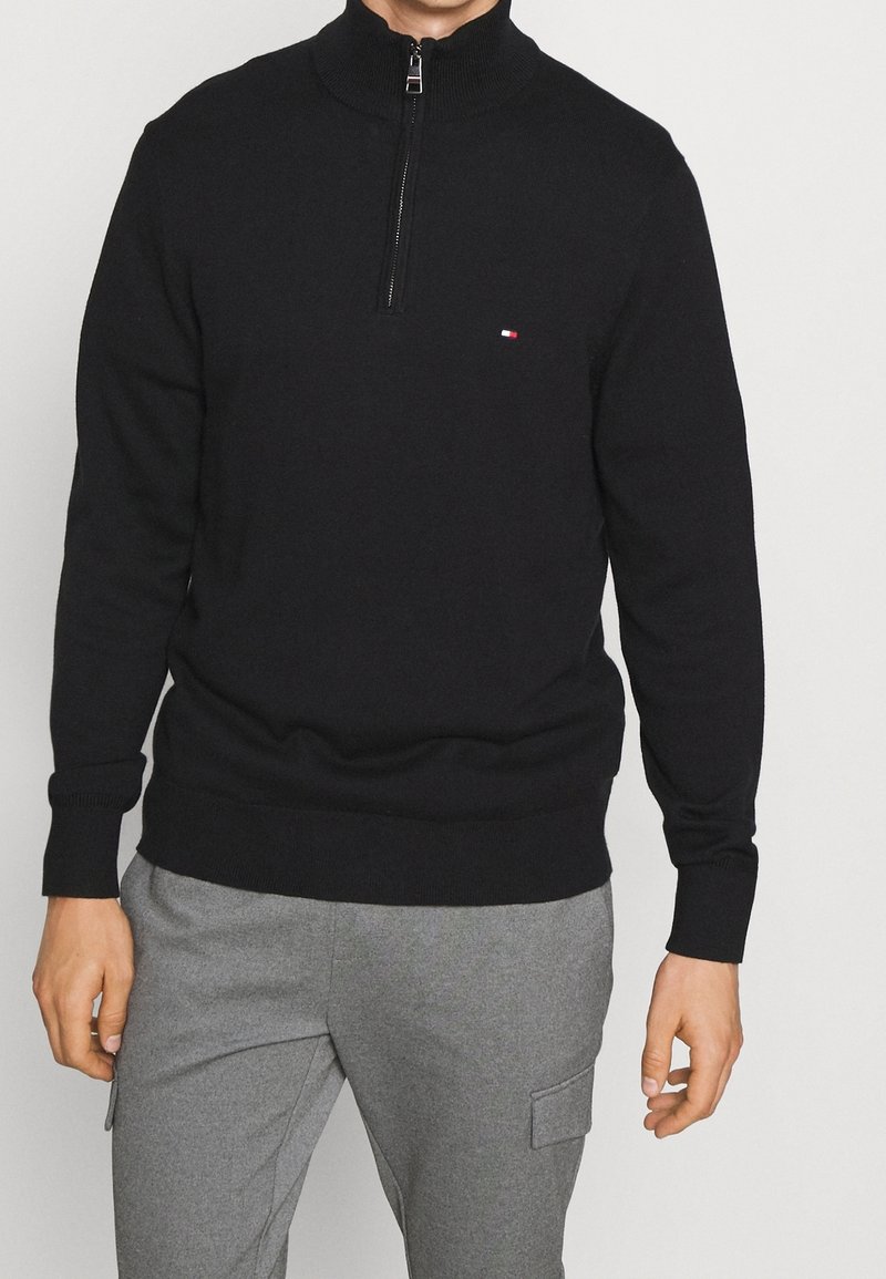 Homme portant un pull noir à fermeture éclair quart de zip avec un petit logo, associé à un pantalon gris doté d'une poche latérale.