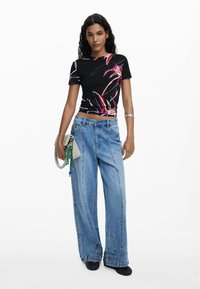 Top corto a maniche corte nero a coste con stampa floreale colorata, abbinato a jeans in denim azzurro chiaro larghi; tiene in mano una borsa grigia con tracolla a catena.