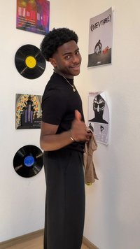 Črna majica s kratkimi rokavi, ohlapne temne hlače, drži svetlo rjavo jakno. Ozadje ima vinile in plakate z različnimi barvami in vzorci.
