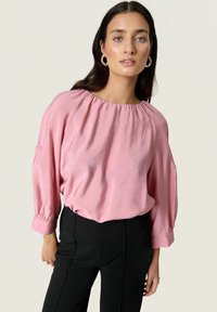 Blusa di raso rosa con scollo arricciato, vestibilità ampia e maniche a palloncino a tre quarti, abbinata a pantaloni neri aderenti.