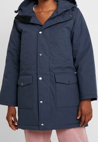 Parka con capucha en azul marino, con botones a presión, dos bolsillos frontales y puños ajustables. Hecha de una tela texturizada para brindar calor y comodidad.