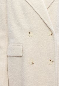 Dvojřadový kabát v krémové barvě, vyrobený z texturované tkaniny. Obsahuje velké světlé knoflíky a límec s výstřihem, s detailem boční kapsy.