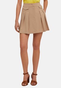 Gonna plissettata khaki con doppie tasche frontali e una texture liscia. Presenta un vitino alto e una silhouette svasata, abbinata a sandali con tacco marroni.