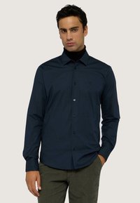 Camicia button-up blu navy in tessuto liscio con maniche lunghe e tasca a sinistra sul petto. Indossata con un dolcevita nero sotto.