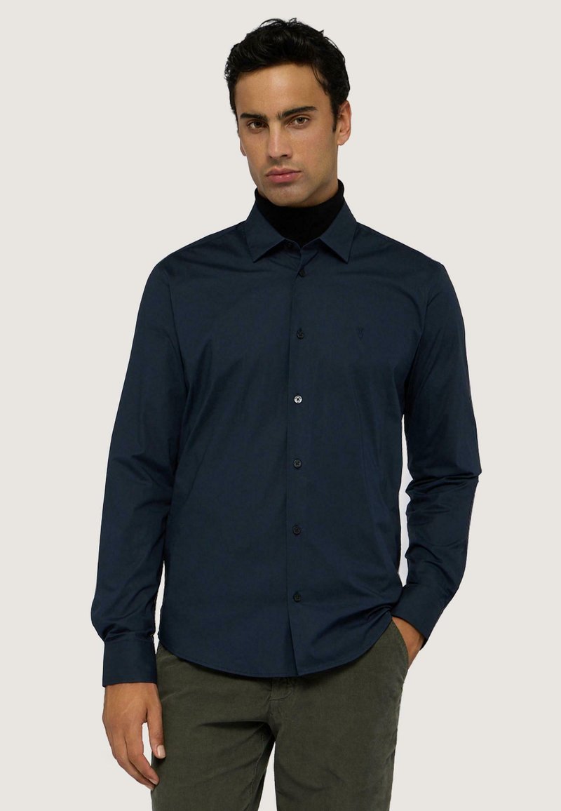 Camicia button-up blu navy in tessuto liscio con maniche lunghe e tasca a sinistra sul petto. Indossata con un dolcevita nero sotto.