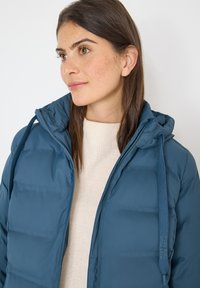 Blå dunjakke med hætte, der har lynlås og snoretræk. Bæres over en let strikket sweater, der viser en glat tekstur.