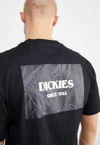 Camiseta de algodón negra con un gran gráfico rectangular gris en la parte posterior que presenta texto blanco que dice "DICKIES SINCE 1922" y un patrón floral.