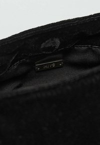 Crna tkanina torbica sa teksturiranom vanjštinom, s glatkom podstavom i malom metalnom oznakom s natpisom "MNG". Unutrašnjost je minimalistička.