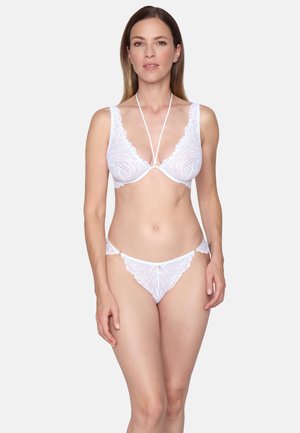 Kvinde iført hvid blonde-bh med halterneck-stropper og matchende blondebikinier, stående mod en ensfarvet baggrund.