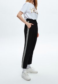 Svarta joggerbyxor med vita sidostripes, elastisk midja och fickor, kombinerade med en vit t-shirt med glänsande guldfärgad text.