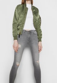 Olivgrön satin bomberjacka med dragkedja, ribbad mudd och en sidoficka, tillsammans med grå skinny jeans med slitningar vid knäna.