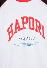 Chemise blanche avec col et manches rouges, texte brodé rouge "HIAPORT" et orthographe phonétique avec un slogan bleu sur la communauté en dessous.