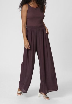 Mujer que lleva una blusa sin mangas de color púrpura oscuro y pantalones anchos a juego con sandalias blancas de tacón, de pie frente a un fondo liso.