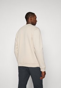 Sweatshirt bege com gola redonda e punhos canelados, apresentando uma textura suave. Usado com calças escuras, mostrado de perfil.