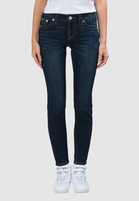 Donkere skinny jeans in blauw met een ontwerp met vijf zakken, contrasterende oranje stiksels en een knoopsluiting, gecombineerd met witte sneakers.