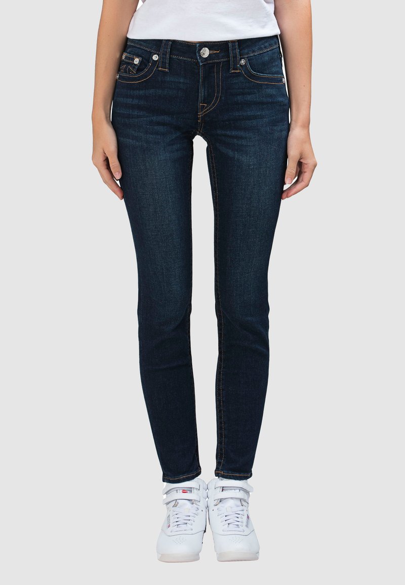 Donkere skinny jeans in blauw met een ontwerp met vijf zakken, contrasterende oranje stiksels en een knoopsluiting, gecombineerd met witte sneakers.