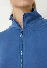 Giacca blu con zip, caratterizzata da un alto colletto e una cerniera decorativa con perline. Il tessuto ha una consistenza liscia e un design aderente.