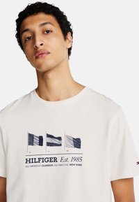 T-shirt bianco in cotone con grafica blu navy di tre bandiere e testo "HILFIGER Est. 1985". Maniche corte e scollatura rotonda.