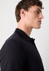 Camisa polo preta com colarinho texturizado, apresentando um fecho de botão e tecido suave. Exibida em um modelo de perfil contra um fundo claro.