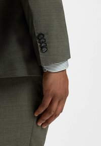 Main d’homme à côté d’une manche de costume vert olive avec quatre boutons noirs et un poignet de chemise rayée visible sous la manche.