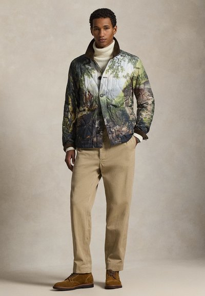 Πουπουλένιο μπουφάν με σχέδιο δάσους σε πράσινο και καφέ, με κουμπιά μπροστά και ψηλό γιακά. Συνδυάζεται με μπεζ παντελόνια και καφέ παπούτσια.
