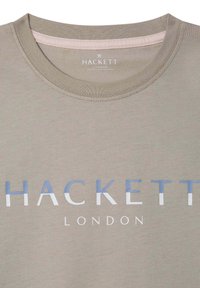 Svijetlo maslinasto zelena majica s okruglim izrezom. Na prsima se nalazi natpis "HACKETT LONDON" ispisan plavim i bijelim slovima. Meka tekstura tkanine.