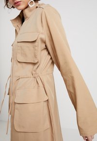 Veste utilitaire beige à manches longues, avec plusieurs poches avant, une taille cintrée avec cordons de serrage, et une fermeture éclair à l'avant.