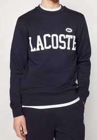 Mörkblå sweatshirt gjord av bomull, har stora vita "LACOSTE"-bokstäver, ribbad mudd och nederkant, med en liten logotyp på bröstet.