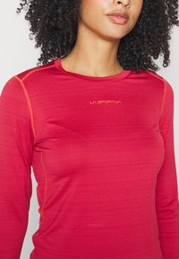 Pitkähihaine urheilu paita kirkkaan punaisesta kankaasta, jossa on heather-mallinen pinta, oranssia ommelta ja pieni logo rinnassa.