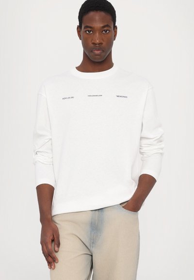 Hvit langermet skjorte med rund hals, med svart tekst på forsiden. Kombinert med lyse beige jeans, hendene i lommene.