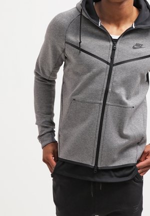 Mężczyzna ubrany w szarą bluzę Nike na zamek z czarnymi lamówkami i kieszeniami typu kangur, w połączeniu z czarnymi spodniami, stoi z rękami przy kieszeniach.