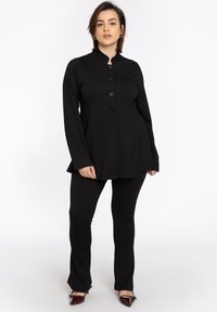 Zwarte button-up jas met hoge kraag en uitlopende zoom, gecombineerd met aansluitende zwarte broek en bordeauxrode puntige schoenen. Gladde stofstructuur.