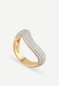Złoty pierścionek z falistym wzorem, ozdobiony wieloma rzędami małych przezroczystych kryształków wzdłuż górnej powierzchni, z logo GUESS wewnątrz.