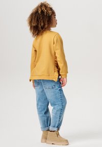 Gul långärmad sweatshirt med sidofåror, ihop med ljusblå baggy jeans och beige ankelstövlar. Lockigt hår synligt från sidan.