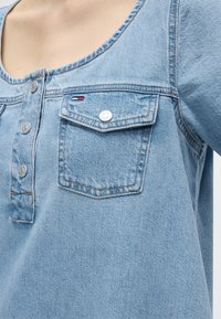 Chemise en denim bleu clair avec boutons-pression argentés et une poche poitrine ornée d’un petit logo rectangulaire rouge, blanc et bleu.