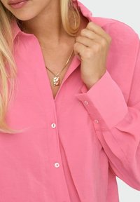 Camicia button-up rosa con una texture morbida, dotata di colletto e dettagli con bottoni. È visibile una collana dorata con un pendente a motivo ondulato.