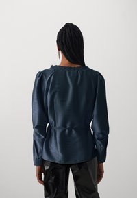 Mørkeblå satengbluse med teksturert stripemønster, rund halslinje og puffede ermer. Har belte i midjen og delikat blonderkant.
