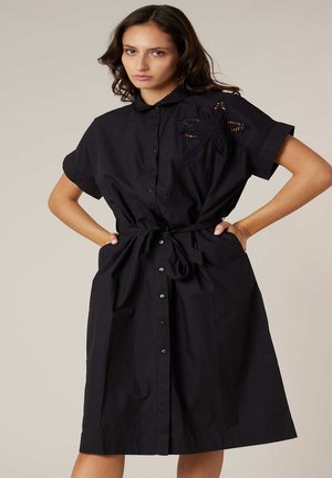 Femme portant une robe noire boutonnée avec des manches courtes, une ceinture à la taille et une broderie florale sur le côté supérieur gauche, debout les mains sur les hanches.