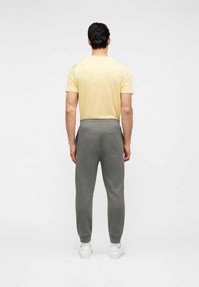 Homme vu de dos portant une chemise à manches courtes jaune pâle, un pantalon de jogging gris avec poches latérales, des chaussettes blanches et des baskets blanches.