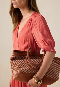 Femme en robe corail tenant un grand sac tissé marron avec des anses tressées et portant un bracelet épais avec des accents dorés.