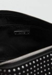 Bolsa negra con cremallera que presenta un exterior liso con detalles de tachuelas metálicas, un interior de tela negra y una pequeña etiqueta de marca en el interior.