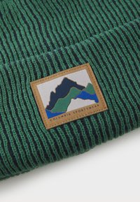 Zöld és tengerészkék bordás kötött sapka, amelyen egy téglalap alakú szőtt folt látható hegygrafikákkal és a "Columbia Sportswear" márkanevével az alján.