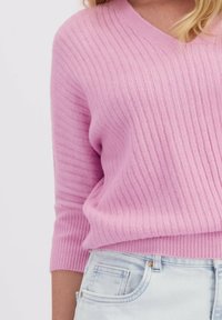 Pull en tricot côtelé rose clair avec un col en V et des manches trois-quarts, associé à un jean en denim bleu clair. Texture douce, design décontracté.