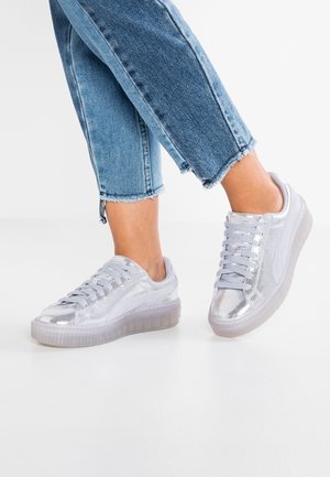 Personne portant un jean court bleu clair et des baskets basses argentées brillantes, debout sur un sol blanc.