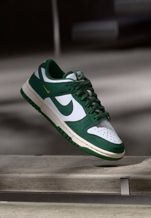 Grøn og hvid Nike-sneaker med gul logo, svævende over en betonoverflade i blødt lys.