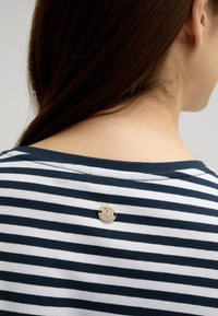 Camicia in cotone a righe blu navy e bianche con un distintivo argentato rotondo sul collo dietro, caratterizzata da una consistenza morbida e una vestibilità casual.
