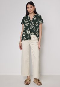 Camicetta a maniche corte di color verde scuro con stampa botanica bianca, abbottonatura frontale, abbinata a pantaloni ampi beige e sandali beige.